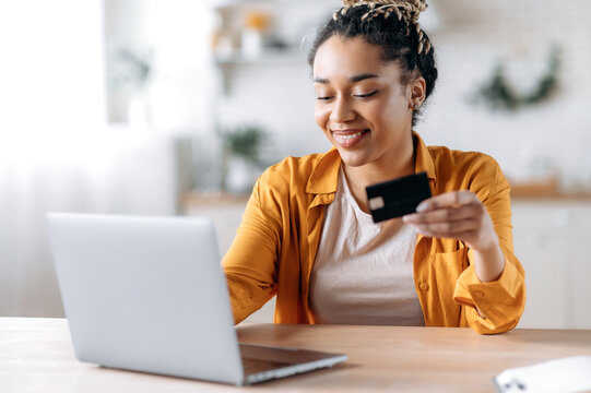 Happy young african american woman stylishly dressed, paying for online purchases, uses a laptop, holding a banking card in hand, typing credit card information on the keyboard and smiling