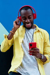 black guy in yellow shirt with red headphones and phone listening to music and laughing standing next to a colorful wall