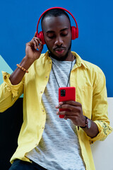 black guy with yellow shirt and red headphones and phone listening to music with his hand on his ears standing next to a colorful wall