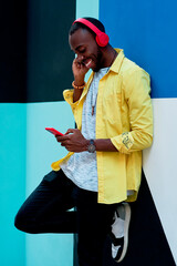 black guy in yellow shirt with red headphones and phone listening to music and laughing standing next to a colorful wall