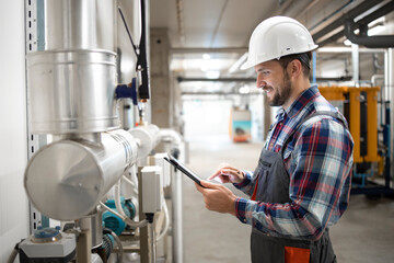 Engineer worker holding tablet computer and setting parameters of heating system in factory boiler room.