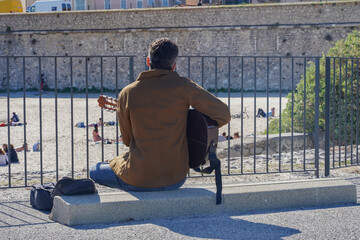 A man sits with his back and plays the guitar near a city beach in Antibes, France.