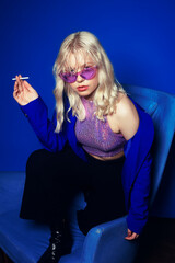 Woman in the style of the 70s, 80s, 90s. A blonde woman in bright glasses and with a cigarette sits on a chair with her legs.  blue jacket, purple glasses, a woman smokes. Grunge blue background