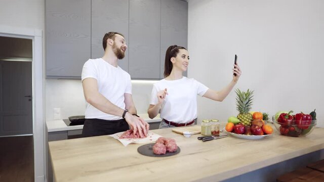 Young cheerful couple cooking food together. They are preparing meat burgers at home in the kitchen. girl has a video conference or chat with a smartphone, saying hi