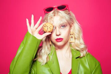 Beautiful blonde in pink glasses and makeup, on a pink background woman in a green raincoat holds a chocolate chip cookie, covering her eye. Model eating sweets, diet, fashion, models grunge