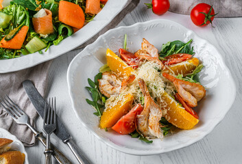 Fresh salad with grilled shrimp, tomatoes, orange, arugula, spinach and parmesan cheese on plate over light wooden background. Healthy food, clean eating, dieting, top view