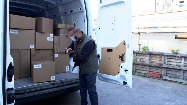 young delivery man scans a QR code from a package with his smartphone and then picks it up and makes a delivery to a customer while wearing a health mask during the global coronavirus pandemic.