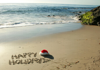 the words "Happy Holidays" written in wet sand with a "Santa Hat" on a secluded beach in "southern california" with the tide of the pacific ocean and blue sky in the background © mike ledray