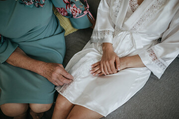 Close-up moment of tenderness between generations — an elderly woman gently touches the edge of a young bride’s satin robe, capturing a quiet connection during a wedding preparation.