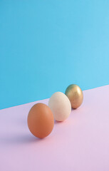 Two fresh eggs and a third painted gold on a light pastel pink and blue background. Minimal Easter creative design.