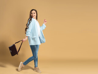 Young beautiful girl with long flowing hair in a white shirt and a blue jacket walks on a pastel orange studio background with a bag in her hand.
