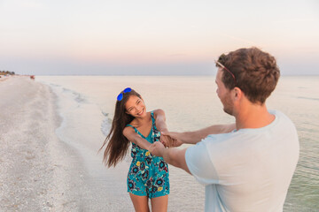 Happy Asian couple of lovers dancing in love on sunset beach. Smiling biracial woman holding hands of boyfriend during honeymoon travel vacation.
