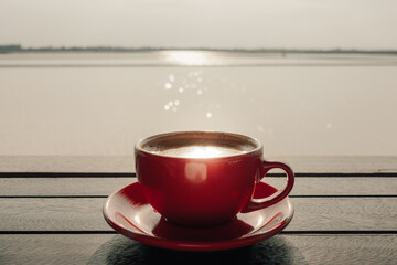 Red cup of hot latte coffee on wooden table at sunrise.