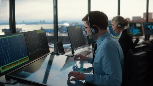 Male Air Traffic Controller with Headset Using Touchscreen Table Panel in Airport Tower. Office Room is Full of Computers with Navigation Screens, Airplane Flight Radar Data for the Team.