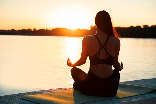 Back view of young woman meditating in sunset , by the river 