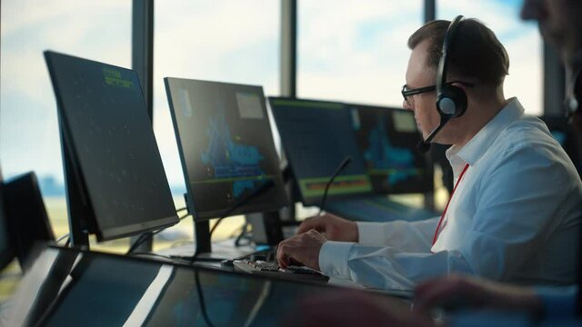 Male Air Traffic Controller with Headset Talk on a Call in Airport Tower. Office Room is Full of Desktop Computer Displays with Navigation Screens, Airplane Flight Radar Data for the Team.
