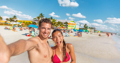 Happy couple taking fun selfie on Florida beach on travel summer vacation. Suntan man and Asian bikini woman in bikini using phone laughing POV. Interracial group portrait on Fort Myers beach holiday. - obrazy, fototapety, plakaty