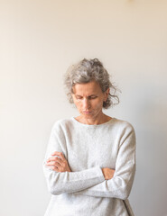 Waist up view of natural middle aged woman looking down with crossed arms against neutral wall background (selective focus)