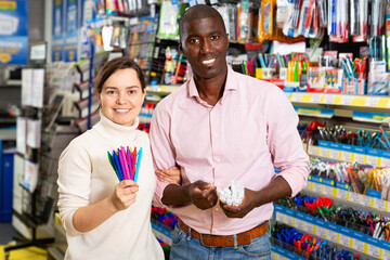 Portrait of glad cheerful smiling Afro American man and Caucasian woman choosing ball pens at stationery store