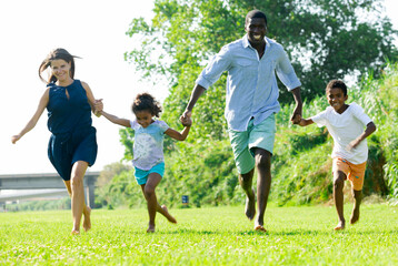 Cheerful mother, father, daughter and son gaily running in summer park during family walk