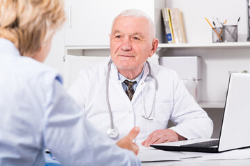 Old female visitor consulting smiling aged man doctor in hospital