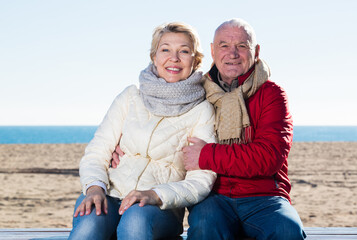 Aged husband and wife sitting together on bench by sea on chilly day