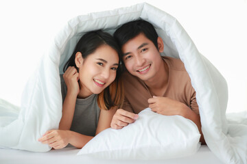 Portrait of cute smiling young Asian lover couple lying on the bed under the comfortable cozy blanket, looking at the camera in the morning at home. Wife and husband relaxing together on the weekend