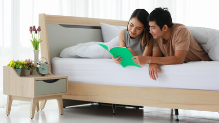 Wide view portrait shot of cute smiling young Asian lover couple lying on elbow under the white blanket on the bed, holding a study book and read it together. Minimalist bedroom interior design