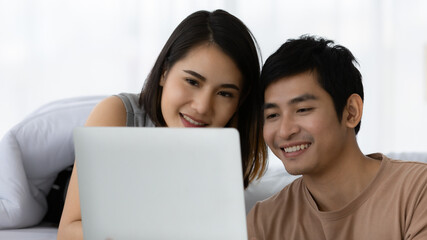 Portrait shot of cute smiling young Asian lover couple watching a movie from modern digital laptop together in the weekends. Husband playing a multiplayer online game with his wife cheering beside