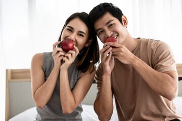 Portrait shot of cute smiling young Asian lover couple sitting on a bed at home, looking at the camera, holding and eating a red apple together in the morning. Concept of fruits and healthy living