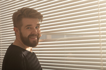 Young man with beard peeks through hole in the window blinds and looks out into the street. Surveillance and curiosity concept