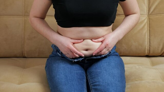 Unrecognizable woman touching belly fat while sitting on the couch.