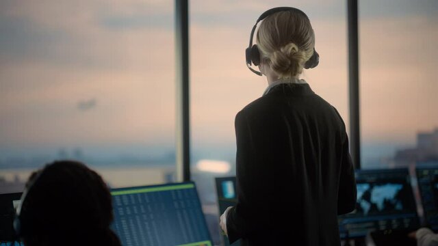 Female Air Traffic Controller with Headset Talk on a Call in Airport Tower. Office Room is Full of Desktop Computer Displays with Navigation Screens, Airplane Departure and Arrival Data for the Team.