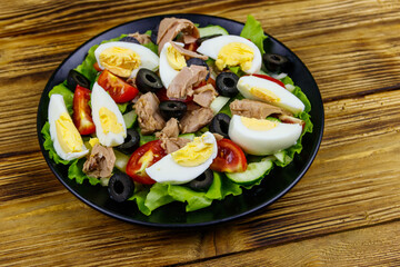 Tasty tuna salad with lettuce, black olives, eggs and fresh vegetables on wooden table
