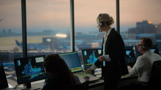 Female Air Traffic Controller with Headset Talk on a Call in Airport Tower. Office Room is Full of Desktop Computer Displays with Navigation Screens, Airplane Departure and Arrival Data for the Team.