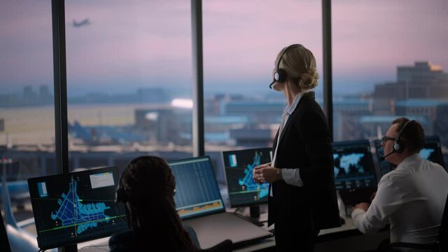 Female Air Traffic Controller with Headset Talk on a Call in Airport Tower. Office Room is Full of Desktop Computer Displays with Navigation Screens, Airplane Flight Radar Data for the Team.