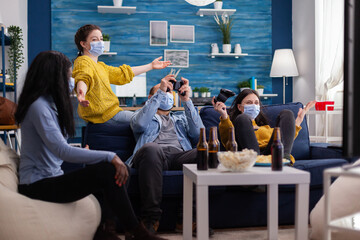 Multiethnic friends celebrating victory playing video games in living room raising hands wearing face mask and keeping social distancing during global pandemic. People enjoying time together