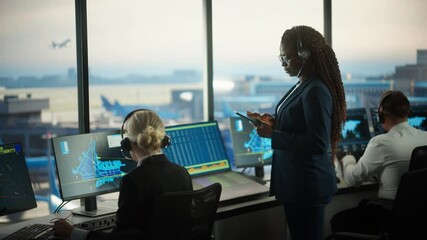 Black Female Air Traffic Controller Holding Tablet in Airport Tower. Office Room is Full of Desktop Computer Displays with Navigation Screens, Airplane Departure and Arrival Data for the Team.