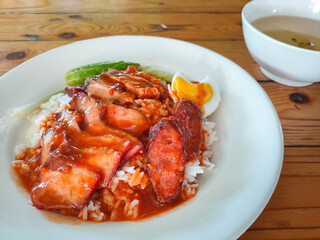 Rice with red barbecue roasted pork, Crispy Pork, and Egg topped with sauce in a white dish with soup on wooden table. It is a delicious menu of Thai. Close up photo.