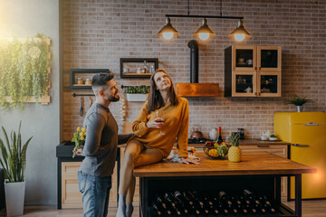 Woman driks wine sitting on table in the kitchen. Man hides flowers behing his back. Bearded man with flowers.