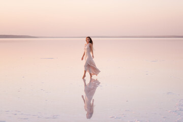 Fototapeta na wymiar Young blonde woman in an evening airy pastel pink, powdery dress stands barefoot on white crystallized salt. Girl with natural make-up, hair is developing. Salt mining trip, walking on water at sunset