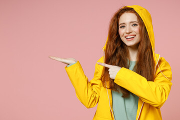 Redhead young woman in yellow waterproof raincoat outerwear hood point finger on empty palm copy...