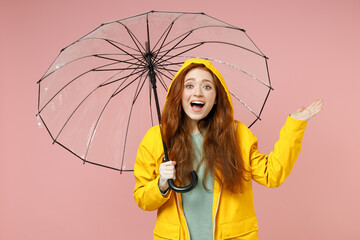 Redhead young fun woman 20s in yellow waterproof raincoat hood outerwear hold transparent umbrella spread hands isolated on pink background studio Outdoors lifestyle wet fall weather season concept.