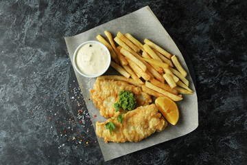 Baking paper with fried fish and chips on black smokey table