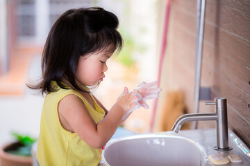 Cute Asian girl washed her hands with soap in the white sink. Children rub the bubbles in their...