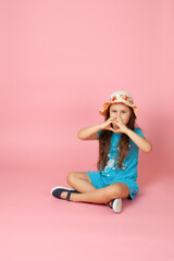 full-length portrait of a girl with long wavy hair in a straw hat and a blue dress sitting cross-legged on the floor with a heart made of fingers, isolated on a pink background.