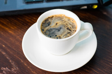 Coffee in a white cup and a laptop on the background on a brown wooden table. Concept of remote work or training, the start of the working day in the office.