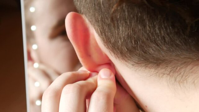 A teenager inserts an earring into his ear while looking in a backlit mirror close-up