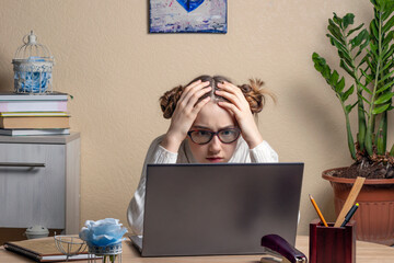 Teenage girl clasping her head in her hands and stares intently at the laptop screen. excited serious teenage girl with glasses is engaged online on a laptop. Online Learning Concept. - obrazy, fototapety, plakaty