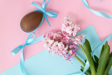 Easter background with chocolate egg with blue bow and pink hyacinth flowers, with blue ribbon on pastel pink and blue colors. Happy Easter concept.